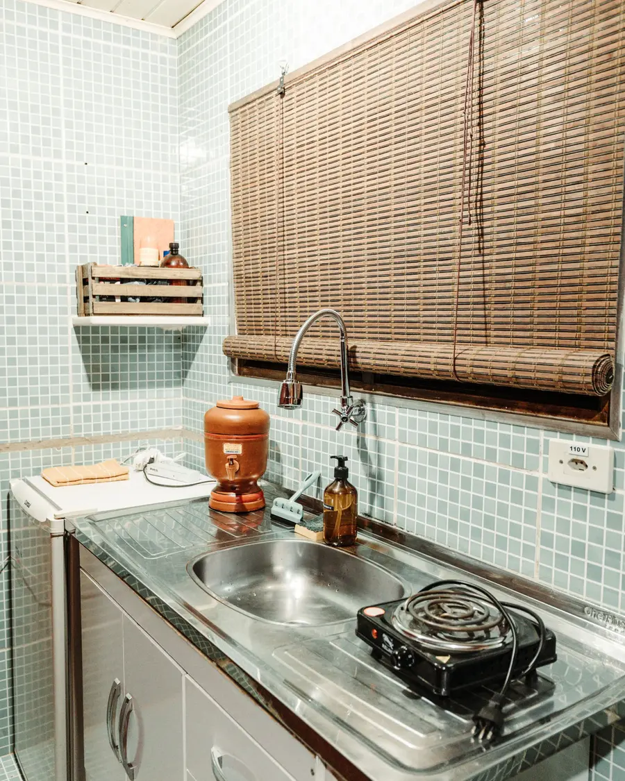 Cozinha pequena de azulejo verde-claro, com pia de inox, fogão e persiana de bambu