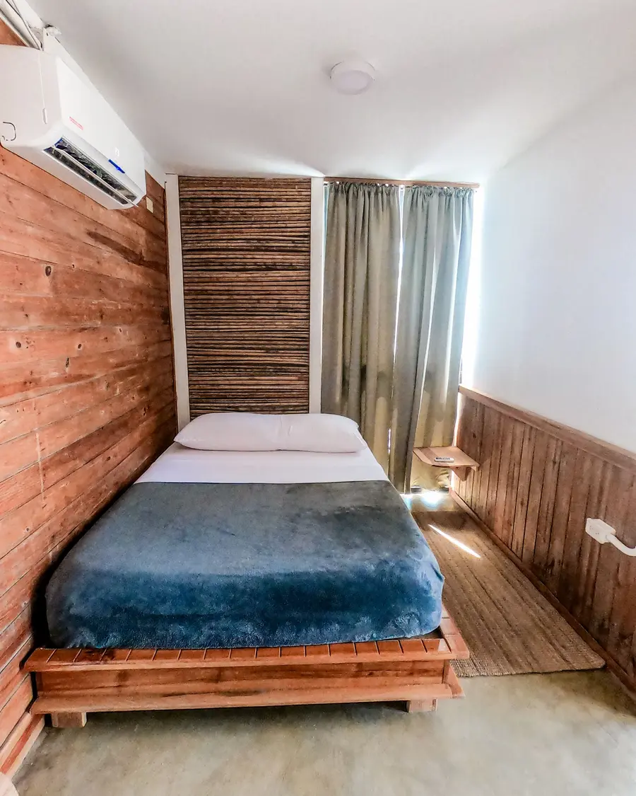 Quarto pequeno com paredes de madeira, cama de casal com manta azul e ar-condicionado